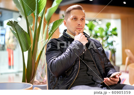 Male in leather jacket with smartphone looking at side while sitting on chair in store against green plant Male in leather jacket with smartphone looking at side while sitting on chair in store against green plant 60905359