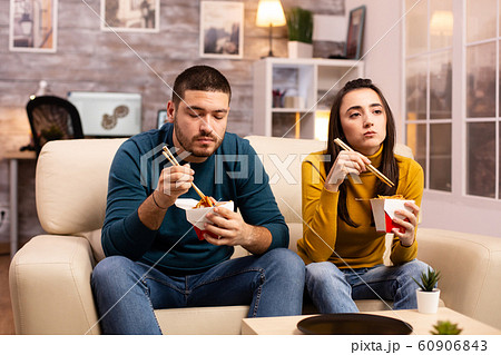 In modern cozy living room couple is enjoying takeaway noodles while watching TV In modern cozy living room couple is enjoying takeaway noodles while watching TV 60906843