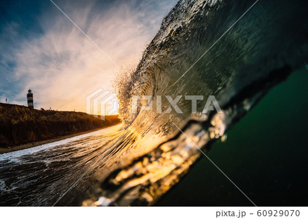 Wave in sea at sunrise. Barrel wave with warm sunrise colors and mountain with lighthouse 60929070