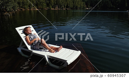 Kid girl lie on a sunbed in sunglasses and a boho silk shawl. Child rest on a flood wood underwater pier. The pavement is covered with water in lake. Kid girl lie on a sunbed in sunglasses and a boho silk shawl. Child rest on a flood wood underwater pier. The pavement is covered with water in lake. 60945307