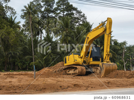 Yellow excavator on a construction site against blue sky. Heavy industry. Close up details of industrial excavator. Large tracked excavator standing on a orange ground with a palms on background. Yellow excavator on a construction site against blue sky. Heavy industry. Close up details of industrial excavator. Large tracked excavator standing on a orange ground with a palms on background. 60968631