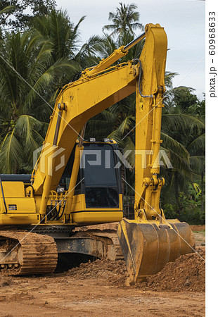 Yellow excavator on a construction site against blue sky. Heavy industry. Close up details of industrial excavator. Large tracked excavator standing on a orange ground with a palms on background. Yellow excavator on a construction site against blue sky. Heavy industry. Close up details of industrial excavator. Large tracked excavator standing on a orange ground with a palms on background. 60968633
