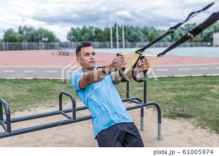 Young sportsman, male athlete, pulls himself on hinges, straps for chest muscles, in summer day in the city, workout fitness, active lifestyle of modern youth, sportswear. Motivation for life. 61005974