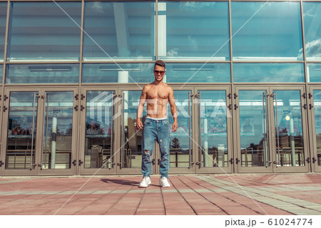 Young guy, athlete dancer, stands background of glass windows, trained happy smiling posing in summer in the city. Hip hop dancing. Jeans sunglasses. 61024774