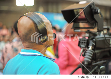 Back portrait of shaved head man with technician works on the broadcasting camera in outdoor event with blurred people in background. 61054343