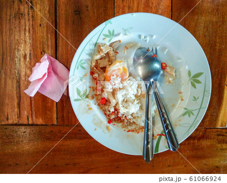 The leftover food on a white plate is steamed rice, fried eggs, pork, spoons and forks, pink tissue paper beside the plate. All laid on a wooden table. The leftover food on a white plate is steamed rice, fried eggs, pork, spoons and forks, pink tissue paper beside the plate. All laid on a wooden table. 61066924