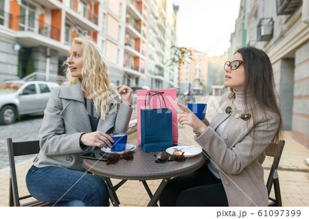 Two young smiling women in an outdoor cafe, drinking coffee, talking, laughing. Urban autumn background 61097399