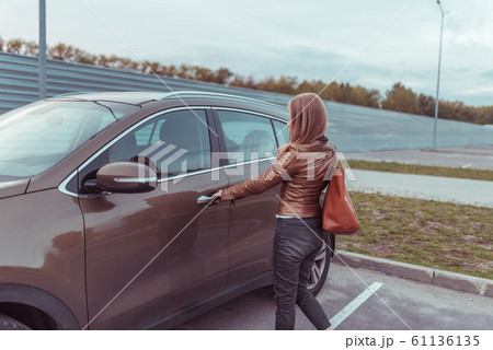 woman opens door crossover SUV in parking lot near shopping center. Autumn day on street in summer, in brown leather jacket, with bag on shoulder. Parking in city, background lawn. woman opens door crossover SUV in parking lot near shopping center. Autumn day on street in summer, in brown leather jacket, with bag on shoulder. Parking in city, background lawn. 61136135