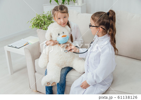Two happy girls playing the game: doctor and patient. They are listening through stethoscope the heartbeat of their patient, teddy-bear. Two happy girls playing the game: doctor and patient. They are listening through stethoscope the heartbeat of their patient, teddy-bear. 61138126