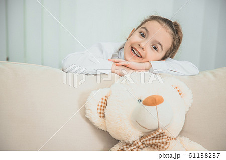Big white soft bear is on the sofa. Girl is standing behind the sofa, leaning over its back, folding her hands, looking at the camera, smiling pleasantly. Big white soft bear is on the sofa. Girl is standing behind the sofa, leaning over its back, folding her hands, looking at the camera, smiling pleasantly. 61138237