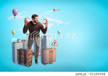 Angry young man in casual clothes holding two alarm clocks with city skyscrapers and hot air balloons on blue background 61164409