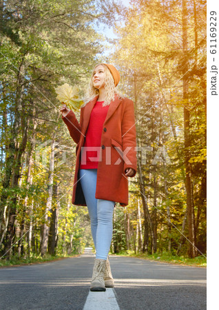 Attractive happy woman in a red coat smiling joyful and blissful holding autumn leaves outside in her hand in a colorful autumn forest walks on asphalt road. 61169229