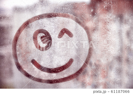 Happy smile face winks painted on window flooded with raindrops on blur red glass background in city 61187066