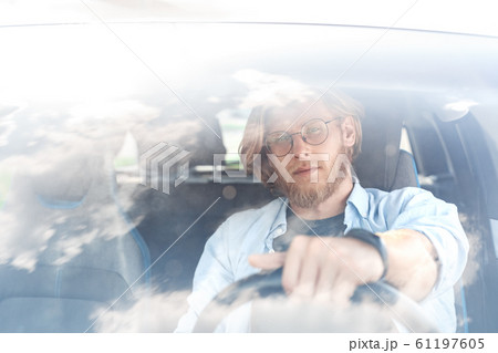 Transportation. Young bearded man in glasses traveling by electric car sitting inside driving looking camera pensive view through windscreen Transportation. Young bearded man in glasses traveling by electric car sitting inside driving looking camera pensive view through windscreen 61197605