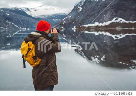 Traveler man with a yellow backpack wearing a red hat standing on the background of mountains and lake makes a photo landscape. Travel lifestyle concept. Shoot from the back 61199232
