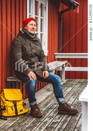 Traveler man with a yellow backpack wearing sits near the wooden red colored house. Travel lifestyle concept. 61199326