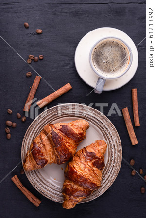 Croissants on a ceramic plate, a cup of coffee, cinnamon sticks and coffee beans on a black wooden table. 61213992