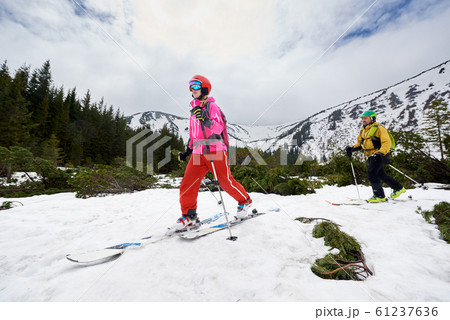 Low angle view of woman walking on ski ahead of man in off-piste ski tour on plain. Mountain, sky, trees on background Low angle view of woman walking on ski ahead of man in off-piste ski tour on plain. Mountain, sky, trees on background 61237636