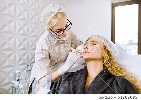Attractive young blond woman is getting a rejuvenating facial injections at modern cosmetology clinic. Young female expert beautician is filling female wrinkles by hyaluronic acid Attractive young blond woman is getting a rejuvenating facial injections at modern cosmetology clinic. Young female expert beautician is filling female wrinkles by hyaluronic acid 61303085