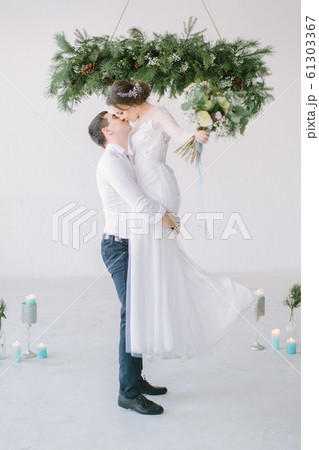 The groom lifted the bride up in his arms. The bride in luxury white wedding dress in the arms of the groom laughing. Wedding ceremony in light white room decorated with pine, flowers and candles. 61303367