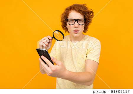 a teenager in glasses with a magnifier in his hand looks at a smartphone in his hand on a yellow 61305117