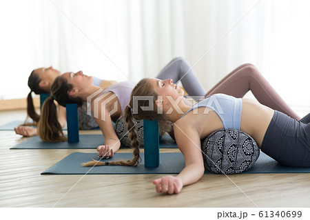 Restorative yoga with a bolster. Group of three young sporty attractive women in yoga studio, lying on bolster cushion, stretching and relaxing during restorative yoga. Healthy active lifestyle Restorative yoga with a bolster. Group of three young sporty attractive women in yoga studio, lying on bolster cushion, stretching and relaxing during restorative yoga. Healthy active lifestyle 61340699