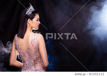 Portrait of Miss Pageant Beauty Contest in Pink sequin Evening Ball Gown with Diamond Crown, Asian Woman points waves hand to side over dark background smoke copy space 61342112