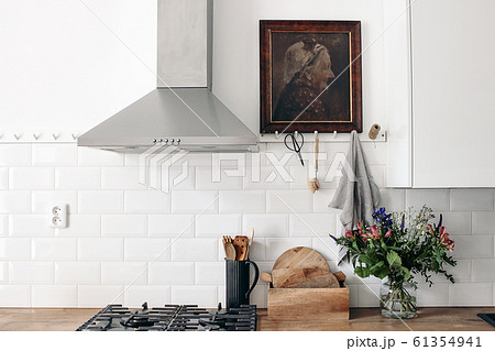Modern eclectic kitchen interior. White brick wall with metro tiles, peg rails and oil painting. Wooden countertop, stainless steel hood and gas stove. Scandinavian design. Home staging, concept. Modern eclectic kitchen interior. White brick wall with metro tiles, peg rails and oil painting. Wooden countertop, stainless steel hood and gas stove. Scandinavian design. Home staging, concept. 61354941