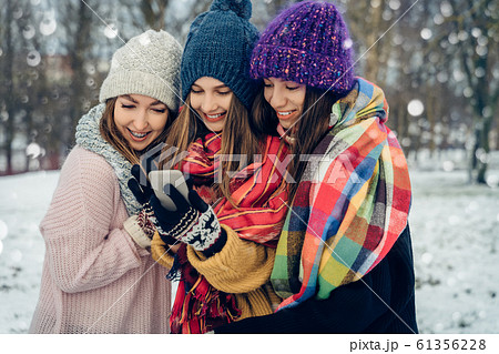 Three women friends outdoors in knitted hats using mobile phone on a snowy cold weather. Group of young female friends outdoors in winter park. Three women friends outdoors in knitted hats using mobile phone on a snowy cold weather. Group of young female friends outdoors in winter park. 61356228