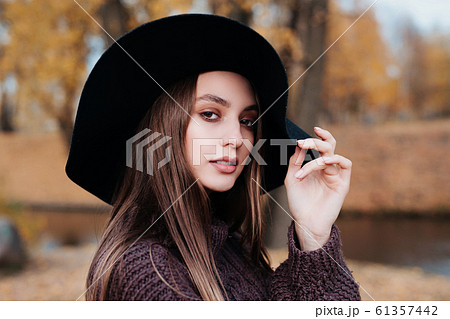 Close-up portrait of a beautiful girl in a black hat standing near colorful autumn leaves. 61357442