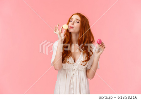 Girl on diet cant resist temptation eating sweets. Alluring funny and cute redhead female holding two tasty macarons want lick dessert, frowning and looking guilty camera, standing pink background 61358216