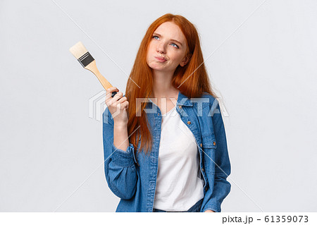 Dreamy and indecisive cute redhead female in denim shirt, smirk and squinting thoughtful looking up imaging something, picturing ideas, holding paintbrush, thinking what color pick to paint 61359073