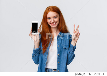 Cute and cheerful alluring redhead female in denim shirt, showing peace gesture smiling and holding smartphone, show app, company website or shopping internet store on mobile screen, white background Cute and cheerful alluring redhead female in denim shirt, showing peace gesture smiling and holding smartphone, show app, company website or shopping internet store on mobile screen, white background 61359101