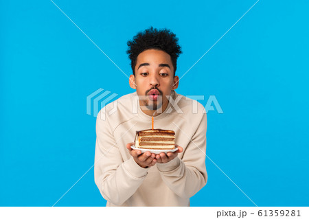 May all wishes fulfill. Cute stylish african-american african american guy with afro haircut, beard, holding birthday cake, blowing candle with folded lips as making wish, standing blue background 61359281
