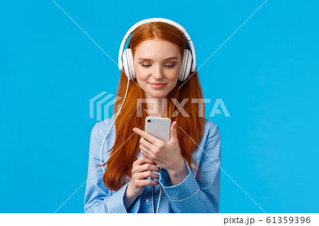 Waist-up portrait good-looking foxy teenage college girl in headphones, listening music, scrolling news feed in morning, checking social media in smartphone, standing blue background in pyjama 61359396