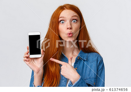 Curious and amused lovely redhead woman with long red hair, folding lips intrigued and excited, discussing new app, photos of classmate with new car, pointing finger smartphone, gossiping 61374656