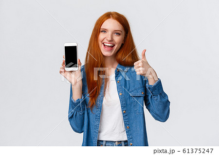 Girl showing friend cool new mobile game. Attractive cheerful redhead woman holding smartphone, introduce application, telephone app, make thumb-up and smiling in approval, recommending 61375247