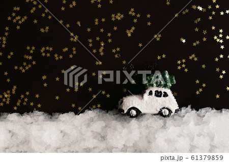 A miniature white toy car carries a Christmas tree on a roof in the snow on a dark background with 61379859