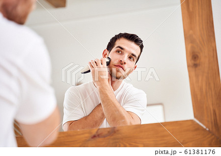 Young male in white T-shirt shaves while standing near mirror in bath 61381176