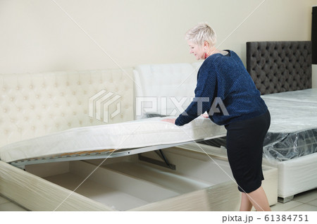An adult stylish woman with a short-cut blonde chooses a large orthopedic bed with a lifting mattress in a furniture store. Selection of quality furniture. choosing a bed for a healthy sleep An adult stylish woman with a short-cut blonde chooses a large orthopedic bed with a lifting mattress in a furniture store. Selection of quality furniture. choosing a bed for a healthy sleep 61384751