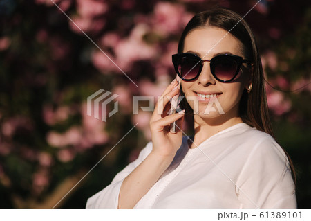 Portrait of attravtive young woman in white shirt and sunglasses use smartphone outside. Speel with someboby by the phone. Background of pink tree 61389101