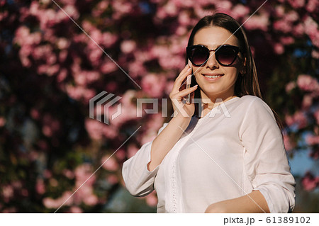 Portrait of attravtive young woman in white shirt and sunglasses use smartphone outside. Speel with someboby by the phone. Background of pink tree 61389102