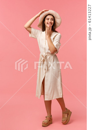 Full-length vertical portrait gorgeous, feminine lovely girl having fun, walking and enjoying sunny day, wearing hat and dress, amused looking right and cover mouth, pink background 61413227