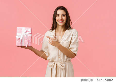 Girl prepared romantic gift for anniversary. Cheerful pretty young woman in cute dress holding gift box and pointing finger at present with delighted, happy smile, standing pink background 61415002
