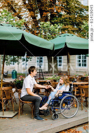 Young pretty woman in wheelchair spending time together with male boyfriend, holding hands, at outdoor cafe in the old city center Young pretty woman in wheelchair spending time together with male boyfriend, holding hands, at outdoor cafe in the old city center 61419045