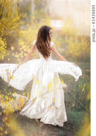 Woman in a white dress in full growth in a garden with trees with yellow flowers 61439895