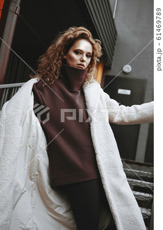 A girl with red curly hair in a white coat poses on the parking stairs A girl with red curly hair in a white coat poses on the parking stairs 61469789