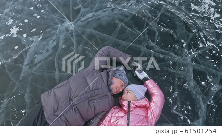 Couple has fun during winter walk against background of ice of frozen lake. Lovers lie on clear ice, have fun, kiss and hug. View from above. 61501254