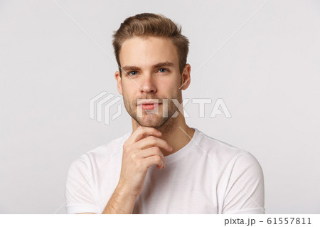 Hmm let me think. Pensive good-looking blond young bearded man in white t-shirt, touching chin and squinting intrigued, thinking, pondering what choice make, standing thoughtful 61557811