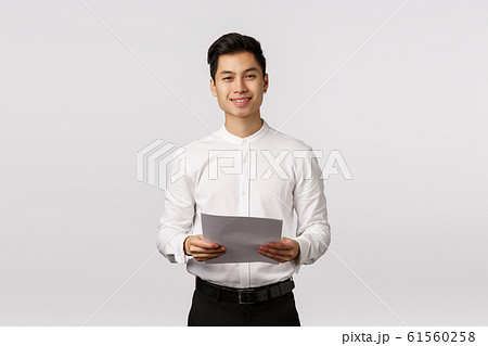 Handsome elegant asian young male coworker, businessman in white collar shirt, black pants, holding papers and smiling camera, successful entrepreneur prepare report, reading speech 61560258